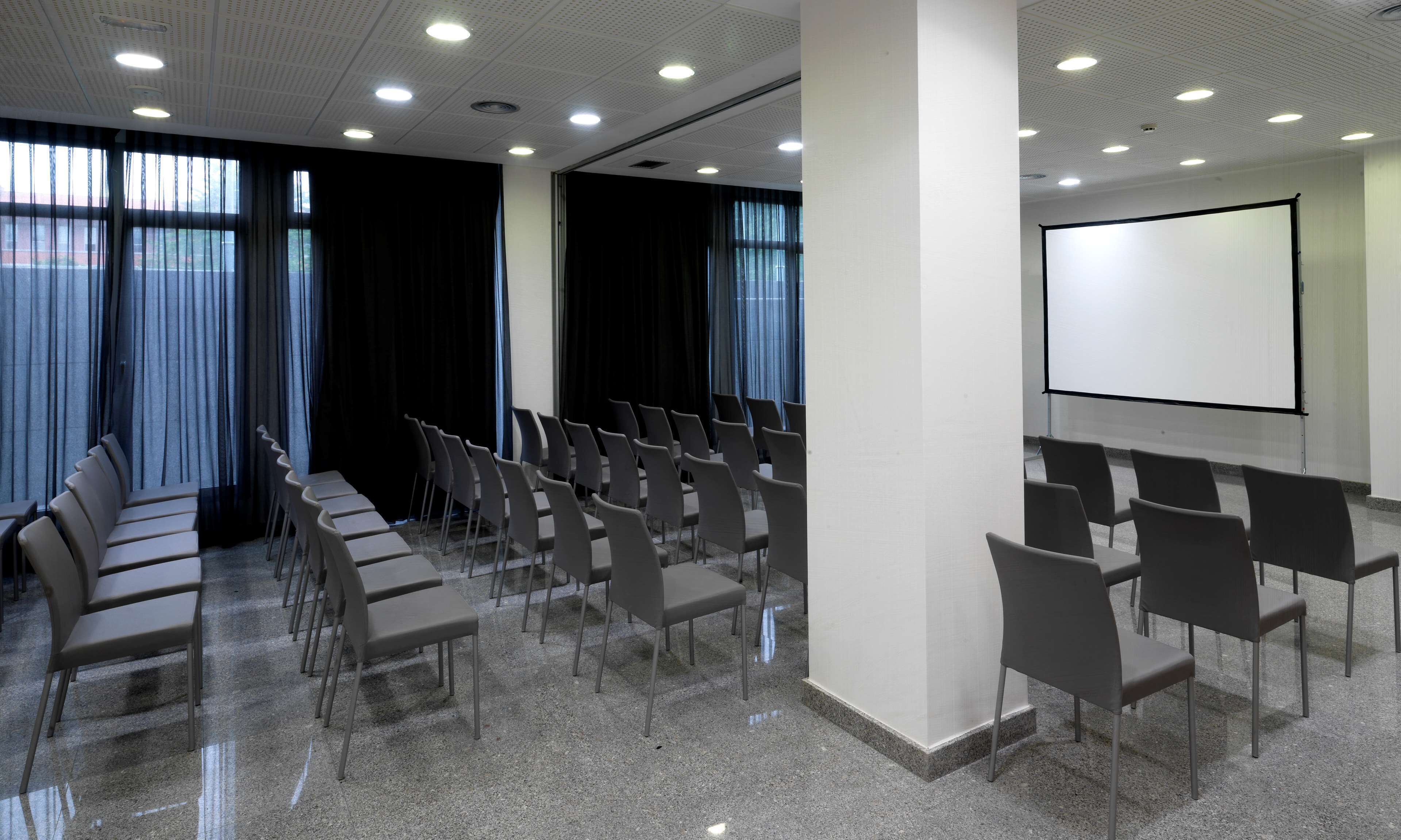 A conference room with rows of chairs, a projector screen, and large windows with curtains.