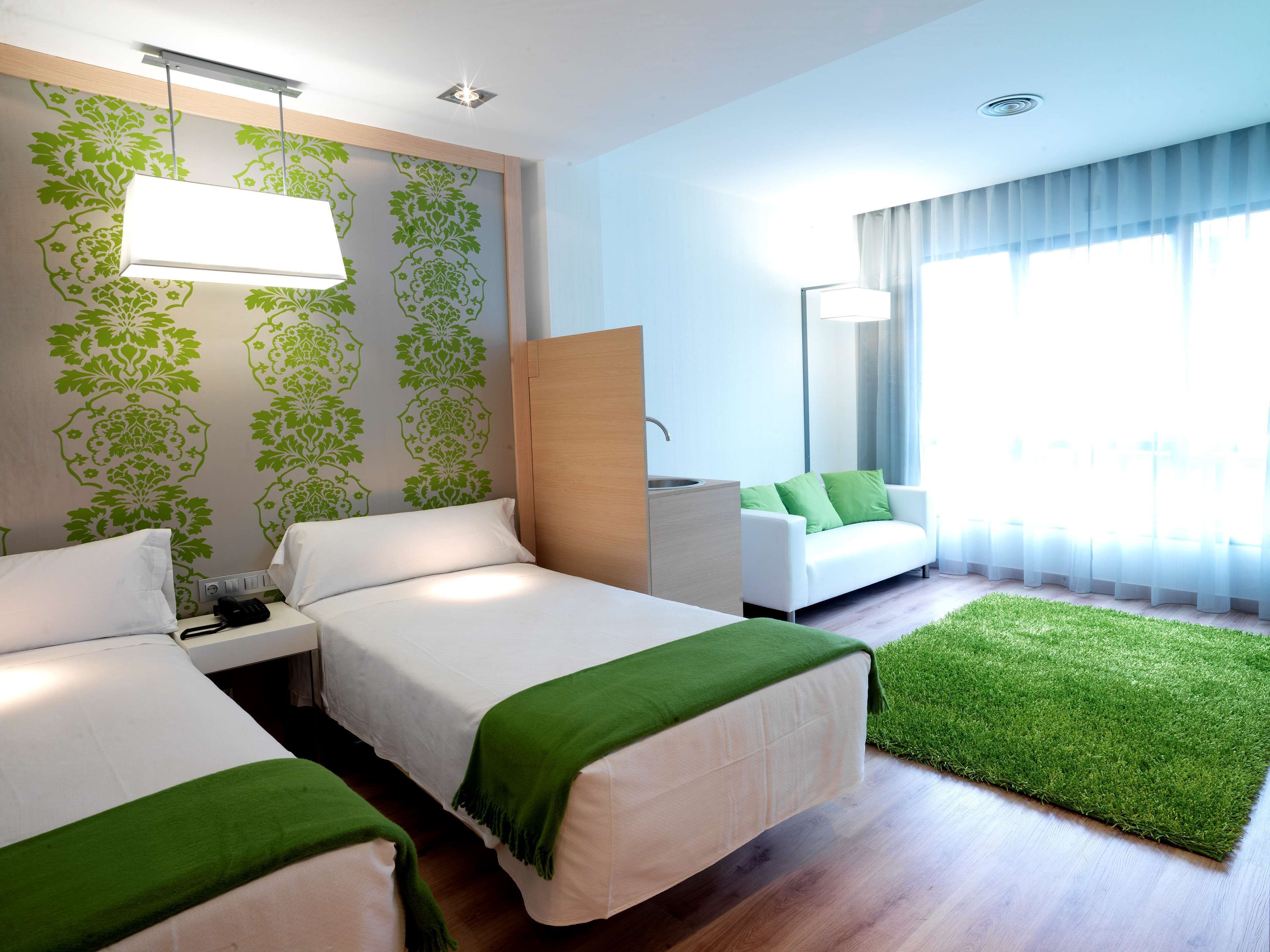 Habitación de hotel con dos camas, un sofá, una mesita, un espejo y una alfombra verde.