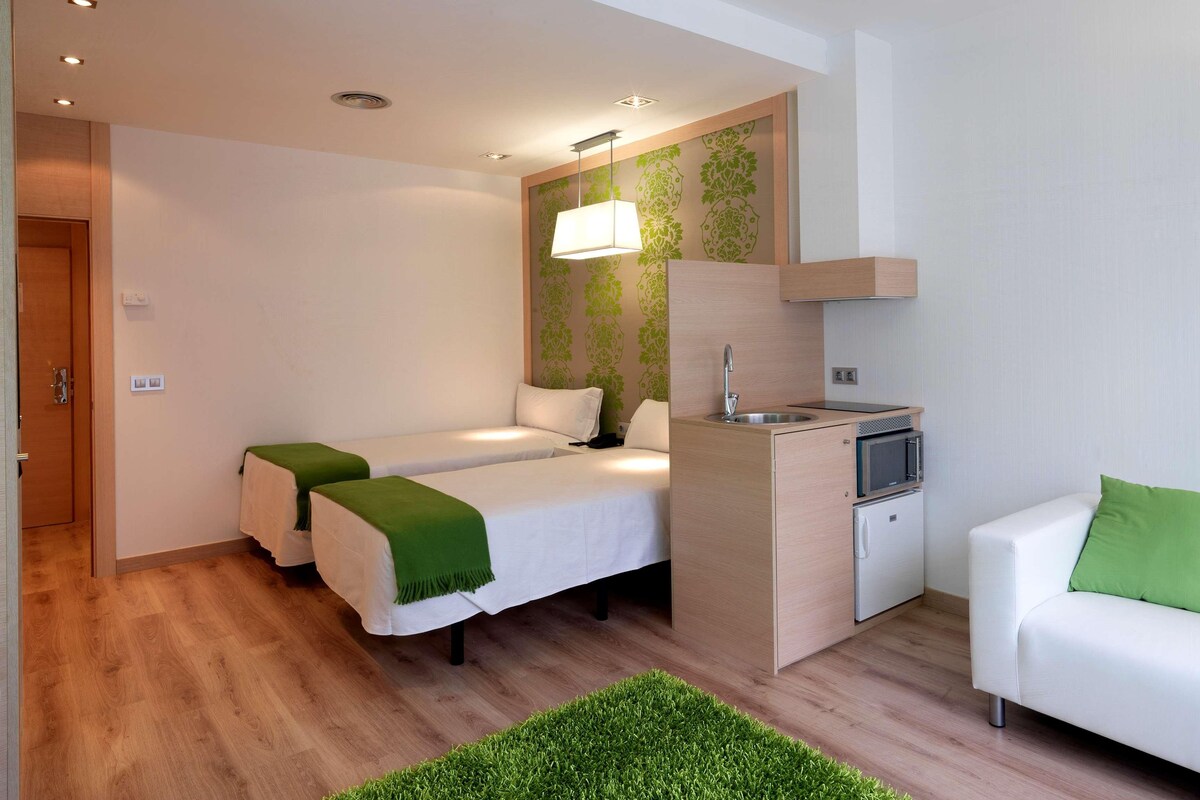 Une chambre d’hôtel compacte avec deux lits simples, une petite kitchenette, un canapé blanc et un mur d’accent vert.