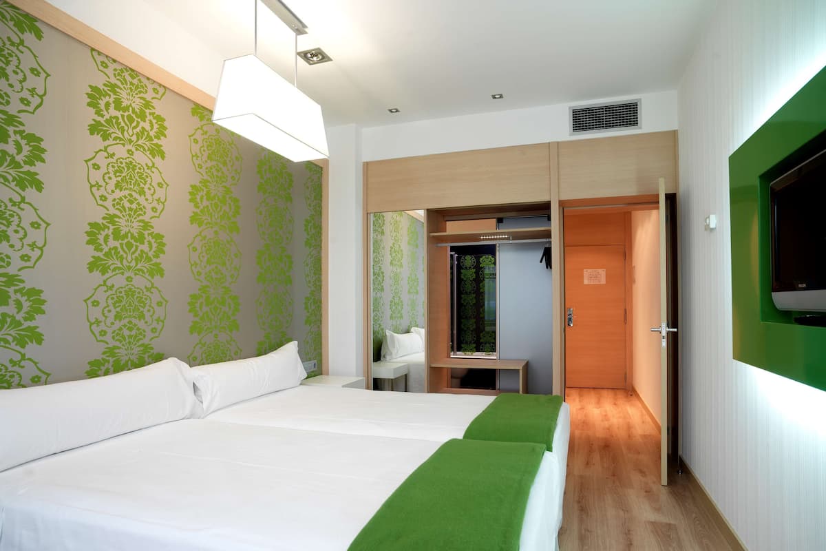 A hotel room with two beds, a green accent wall, a wooden wardrobe, a desk, and a television mounted on the wall.