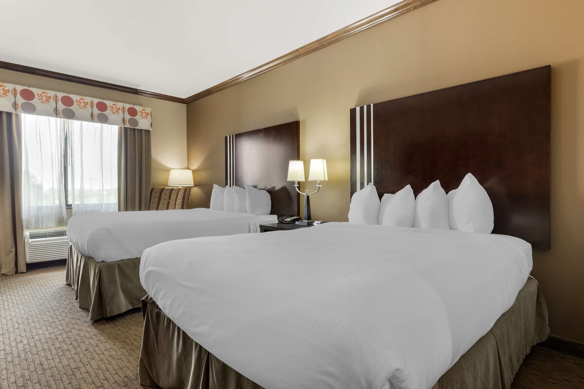 A hotel room with two beds, a large dark headboard, a window with curtains, and a lamp on a nightstand.