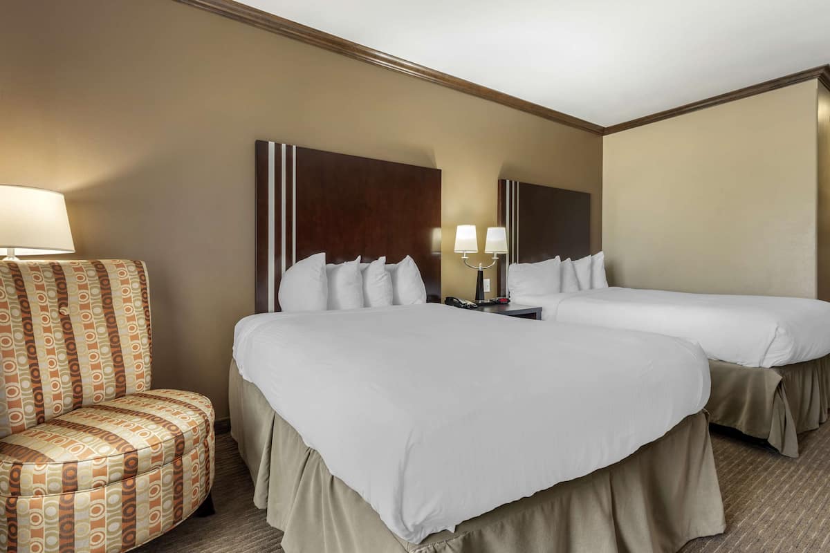 A hotel room with two beds, a patterned armchair, and a nightstand with lamps.