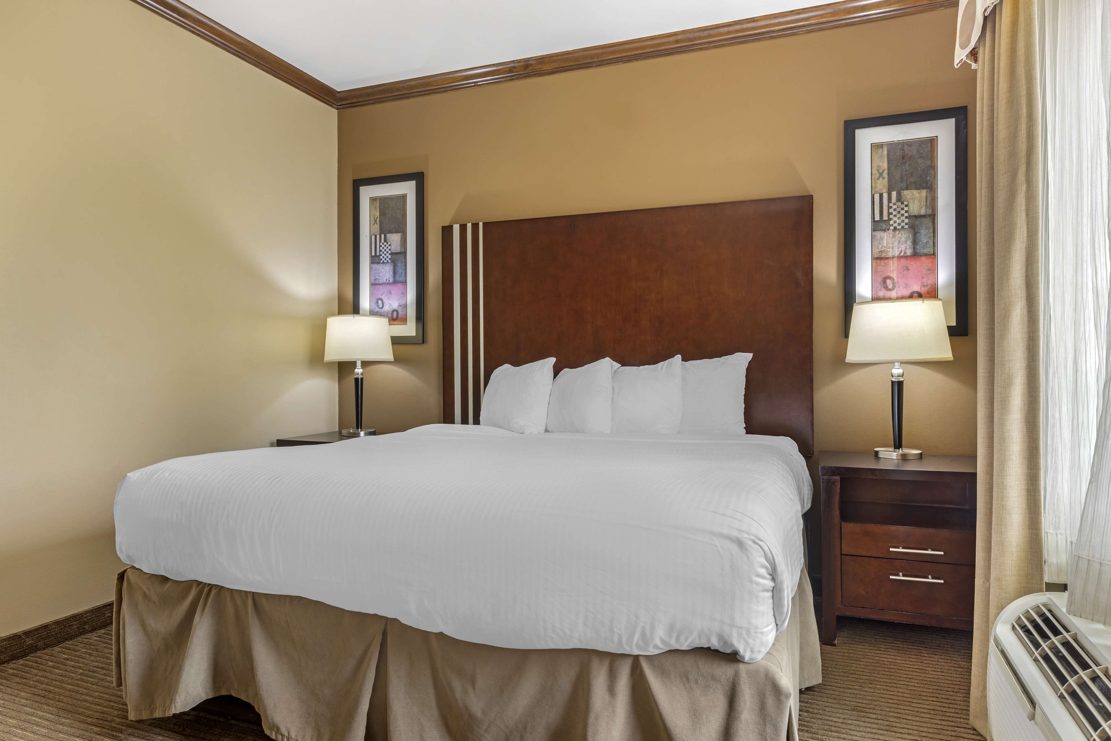 A hotel room with a large bed, two bedside tables with lamps, a wooden headboard, and a framed artwork on the wall.