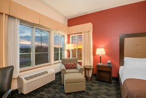 Une chambre d’hôtel avec un lit, une chaise avec un coussin, une table de chevet avec une lampe, un radiateur et une fenêtre avec des rideaux.