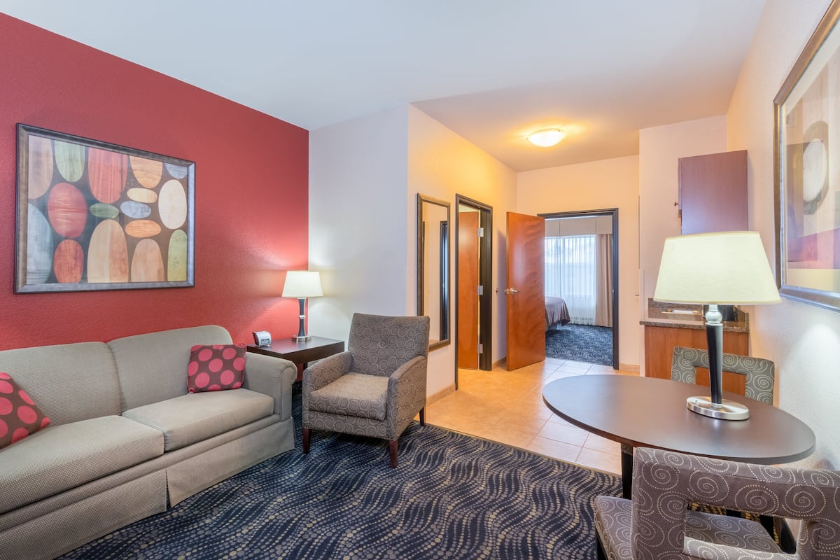 A hotel room with a sofa, armchair, and table with lamps. There is a door leading to another room.