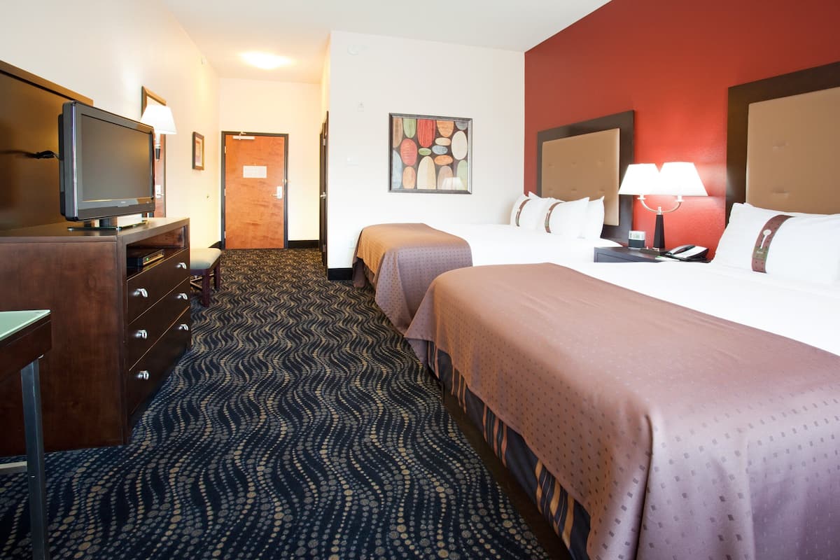 A hotel room with a television, a desk, a dresser, two beds, a carpet, a painting, and a door.