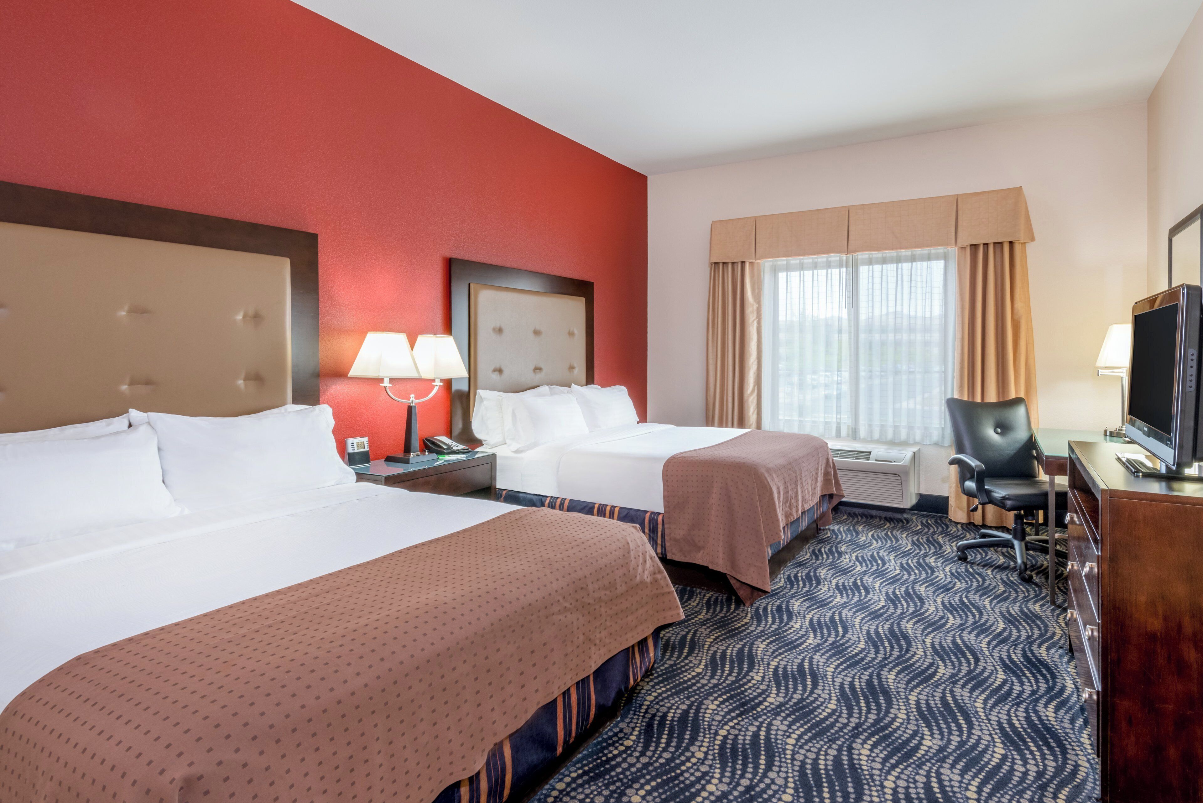 A hotel room with two beds, a desk with a television, a chair, and a window with curtains.
