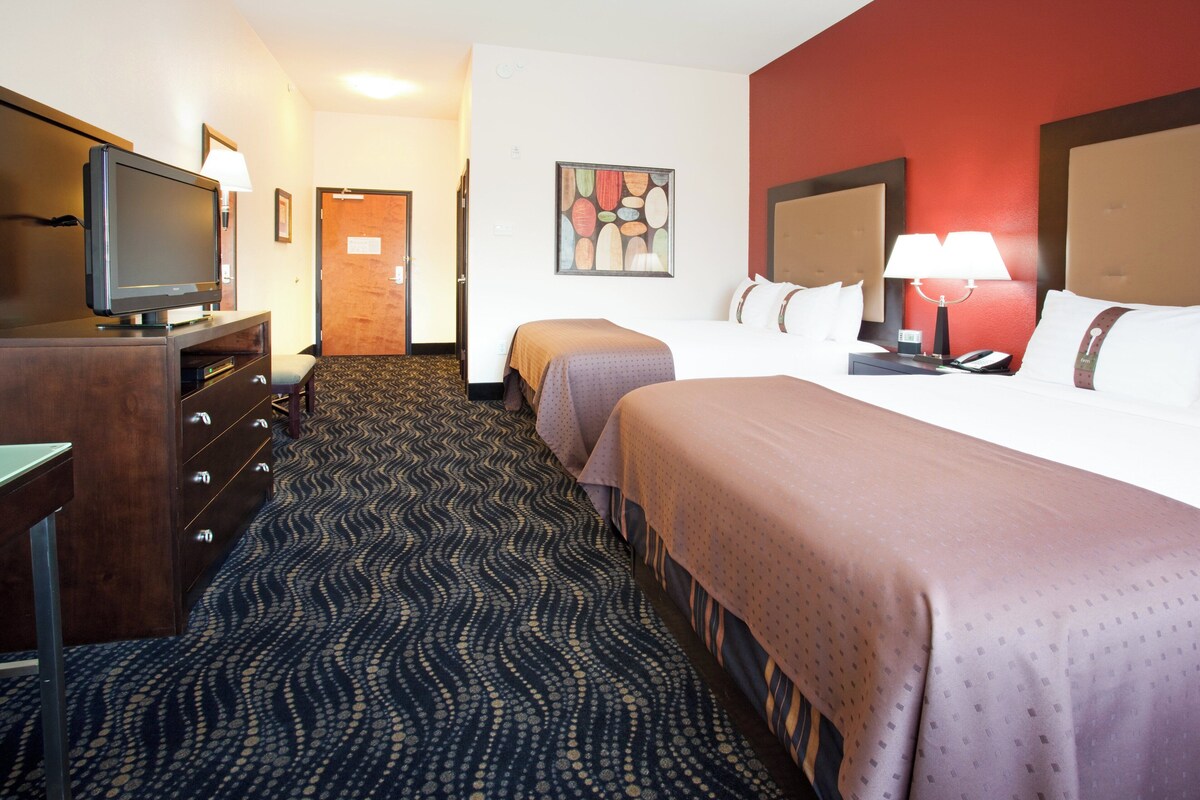 Une chambre d’hôtel équipée d’une télévision, d’un bureau, d’une commode, de deux lits, d’une moquette à motifs et d’un mur d’accent rouge.