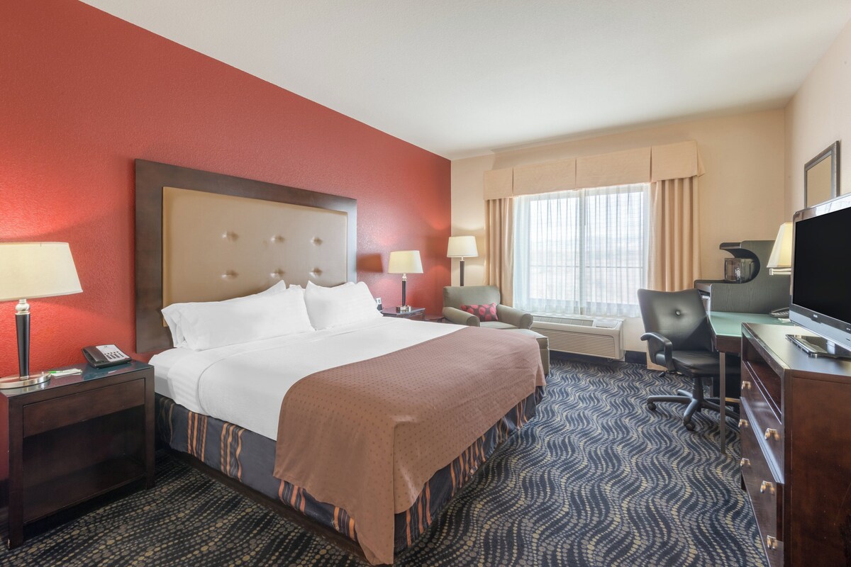 Une chambre d’hôtel avec un grand lit, un bureau avec un ordinateur, une chaise, une télévision et une fenêtre avec des rideaux.