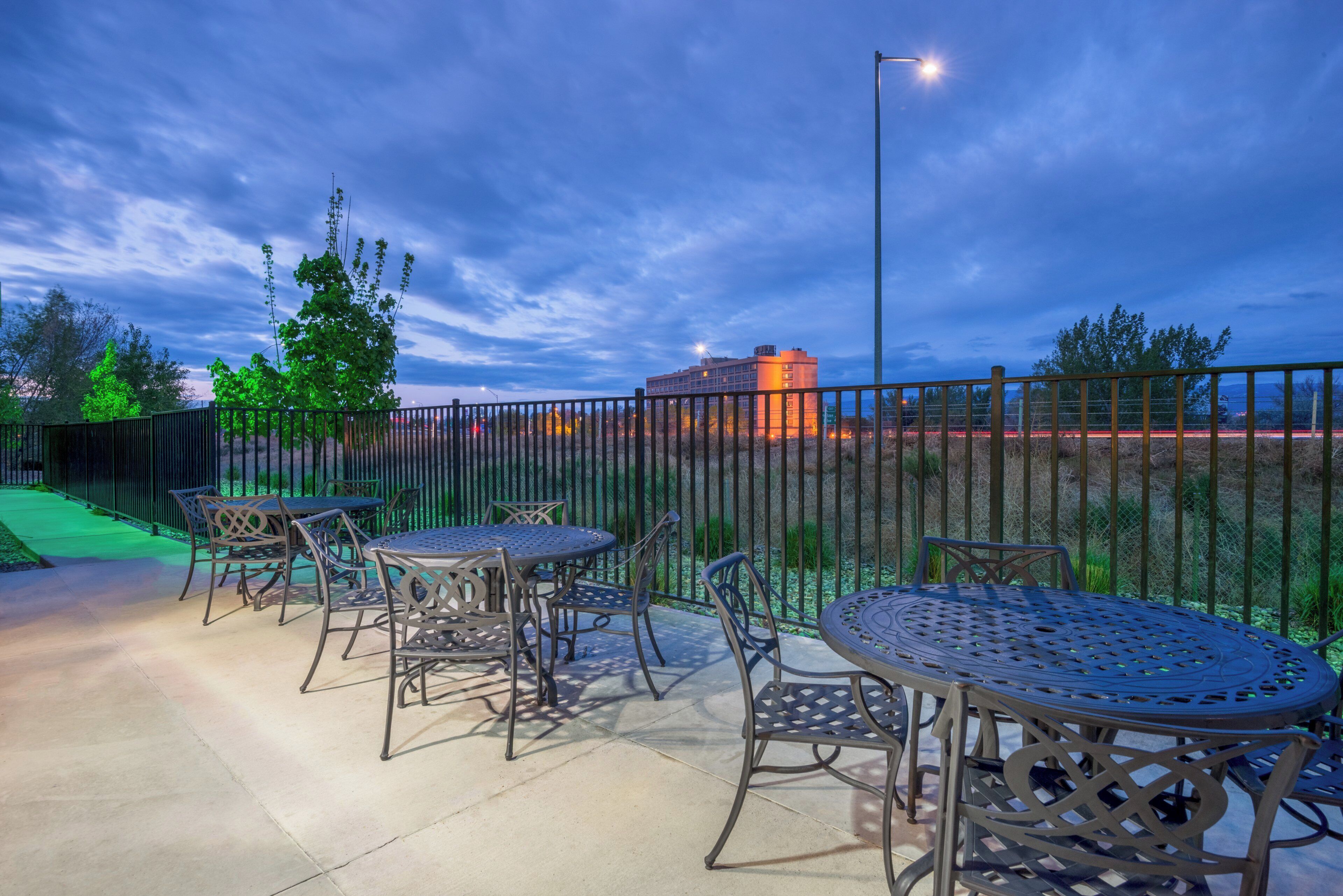 Un patio al aire libre con mesas y sillas de metal, un sendero verde y una vista de un edificio al fondo.
