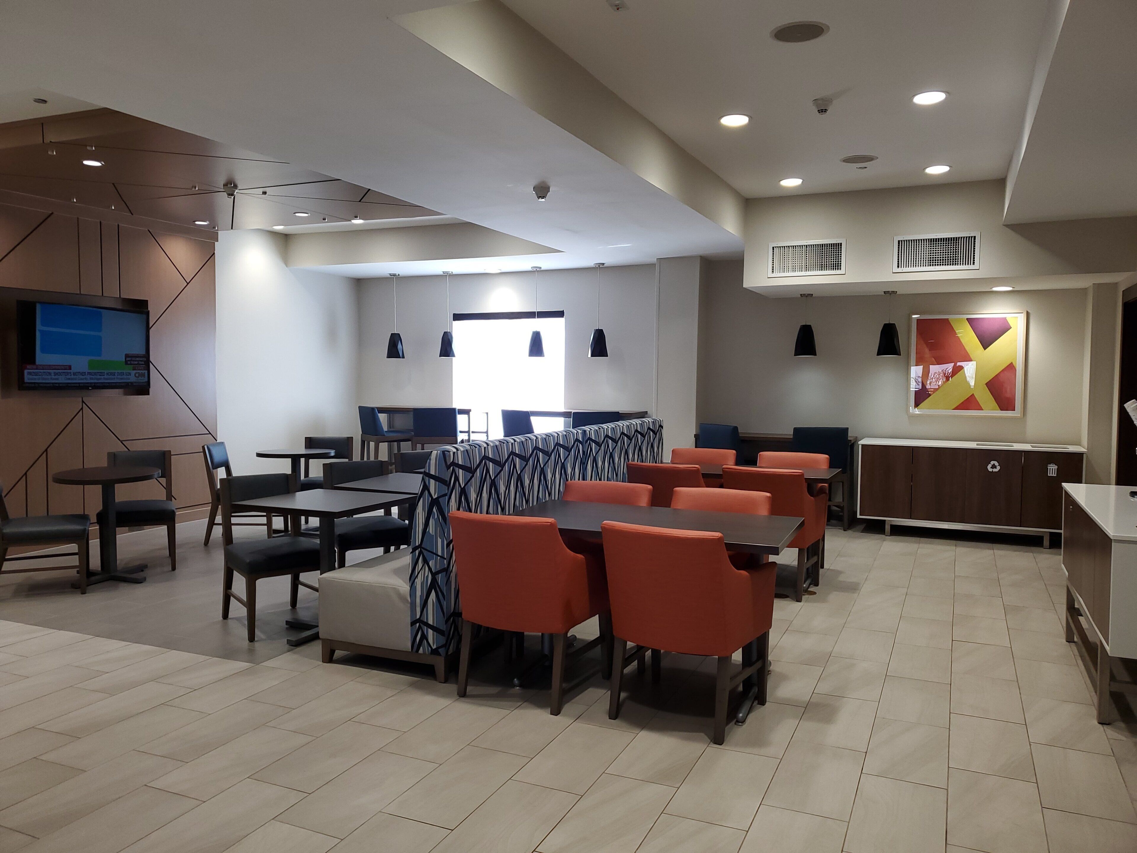 A modern hotel dining area with multiple tables and chairs, a television mounted on the wall, and a large window providing natural light.