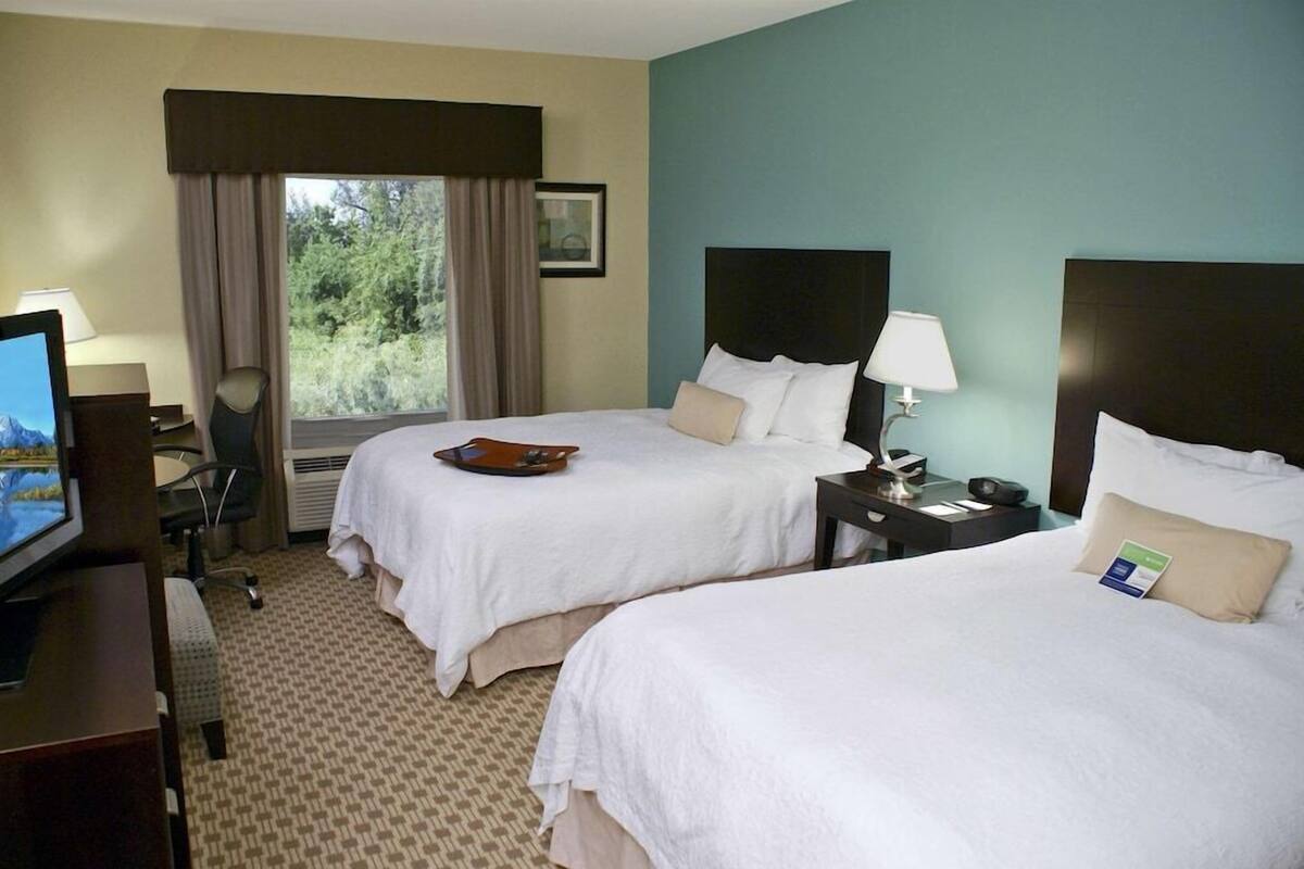 A hotel room with two beds, a desk with a computer, a chair, and a window with curtains.