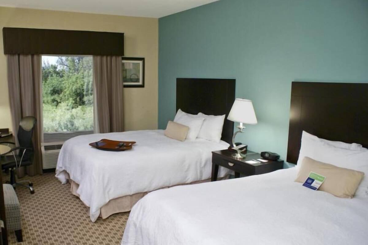 A hotel room with two beds, a desk, a chair, a window with curtains, and a nightstand with a lamp.