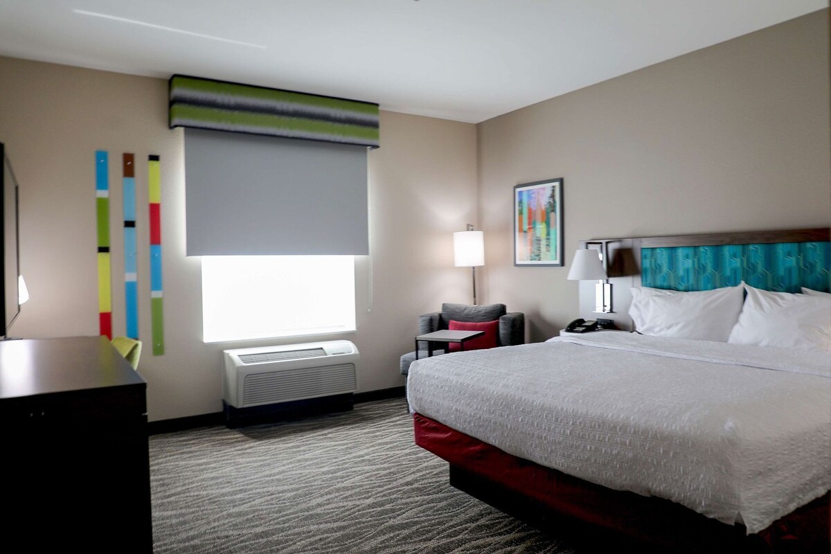 A hotel room with a bed, bedside table, lamp, air conditioning unit, and a window with blinds.