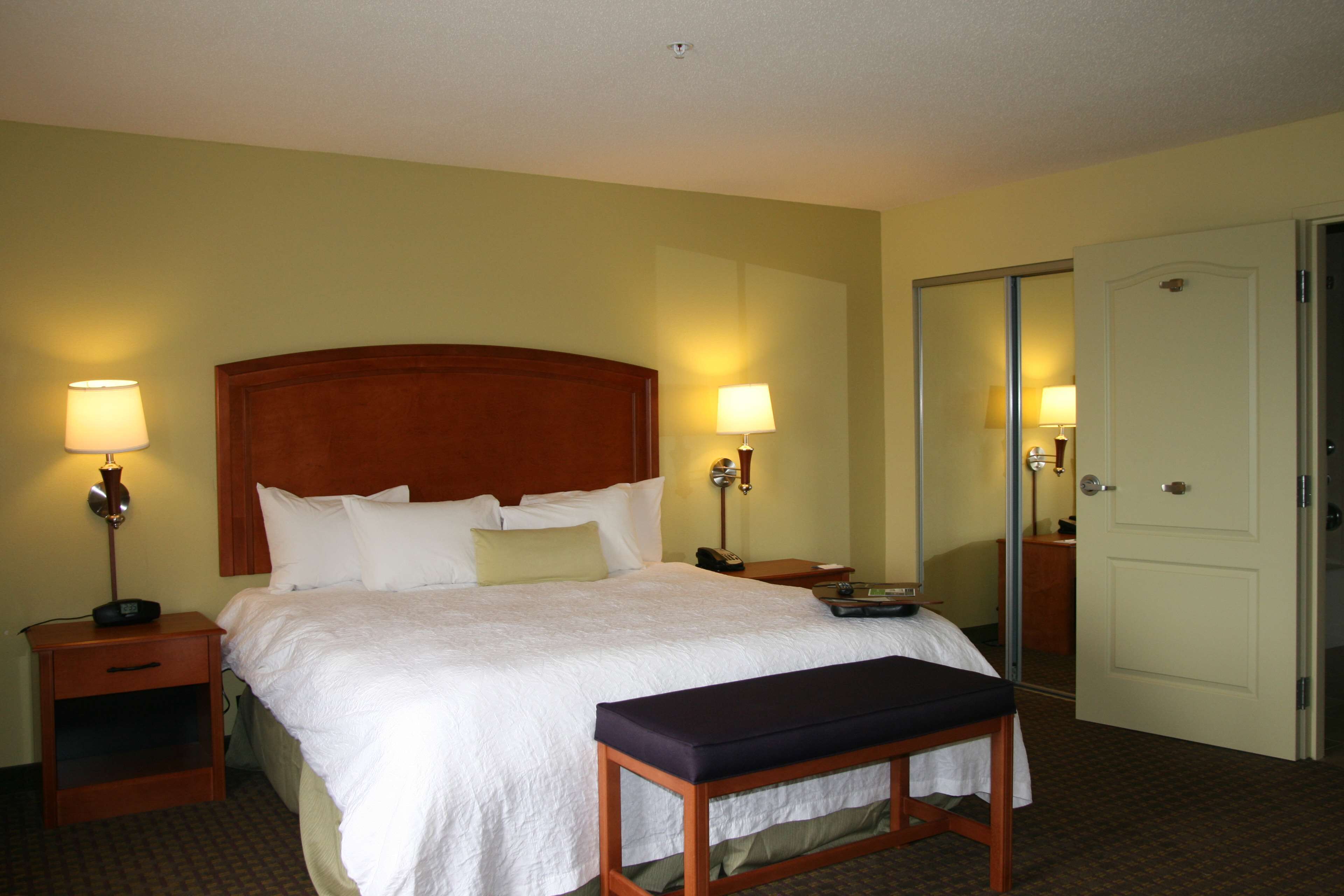 A hotel room with a large bed, two bedside tables with lamps, a bench, and a door.