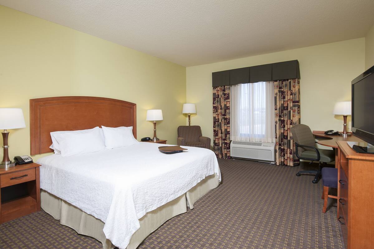 A hotel room with a large bed, two bedside tables with lamps, a desk with a chair, a television, and a window with curtains.