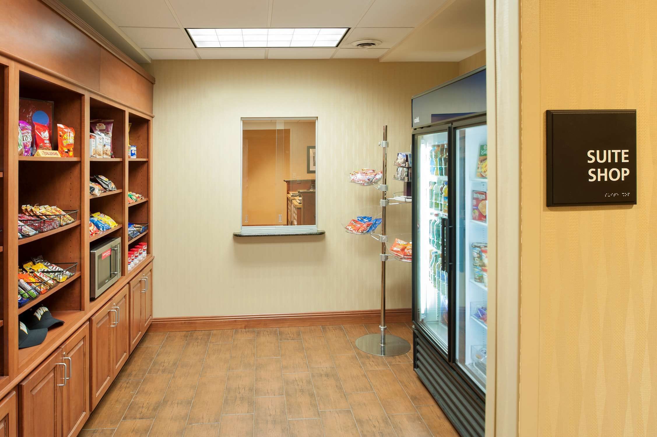 A hotel corridor with a kitchenette on the left, a mirror on the wall, and a refrigerated display case labeled "SUITE SHOP" on the right.