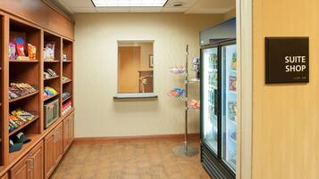 A hotel corridor with a kitchenette on the left, a mirror on the wall, and a refrigerated display case labeled "SUITE SHOP" on the right.