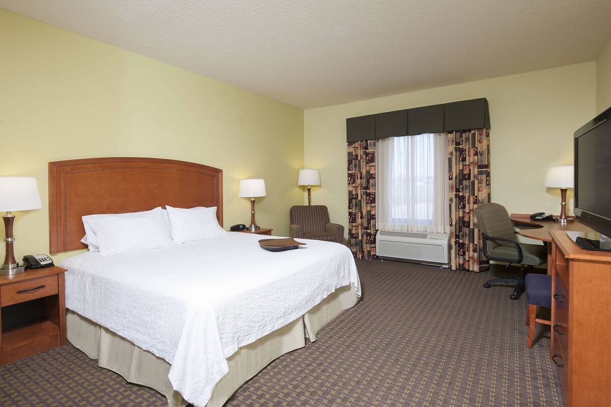 A hotel room with a large bed, two bedside tables, a desk with a chair, a television, and a window with curtains.