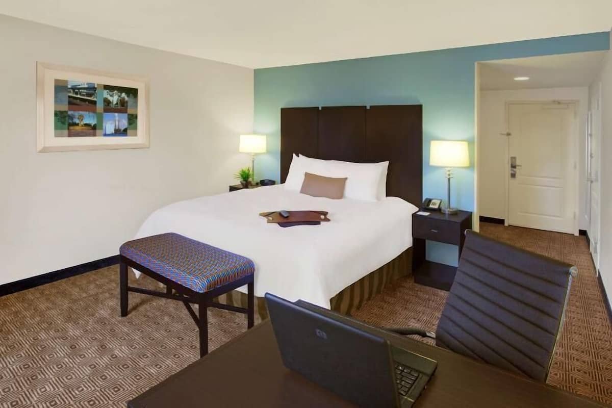 A hotel room with a bed, a desk with a laptop, a chair, a bedside table with a lamp, and a wall-mounted picture.