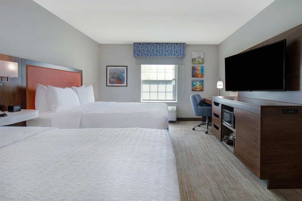A hotel room with two beds, a desk, a television, and artwork on the walls.
