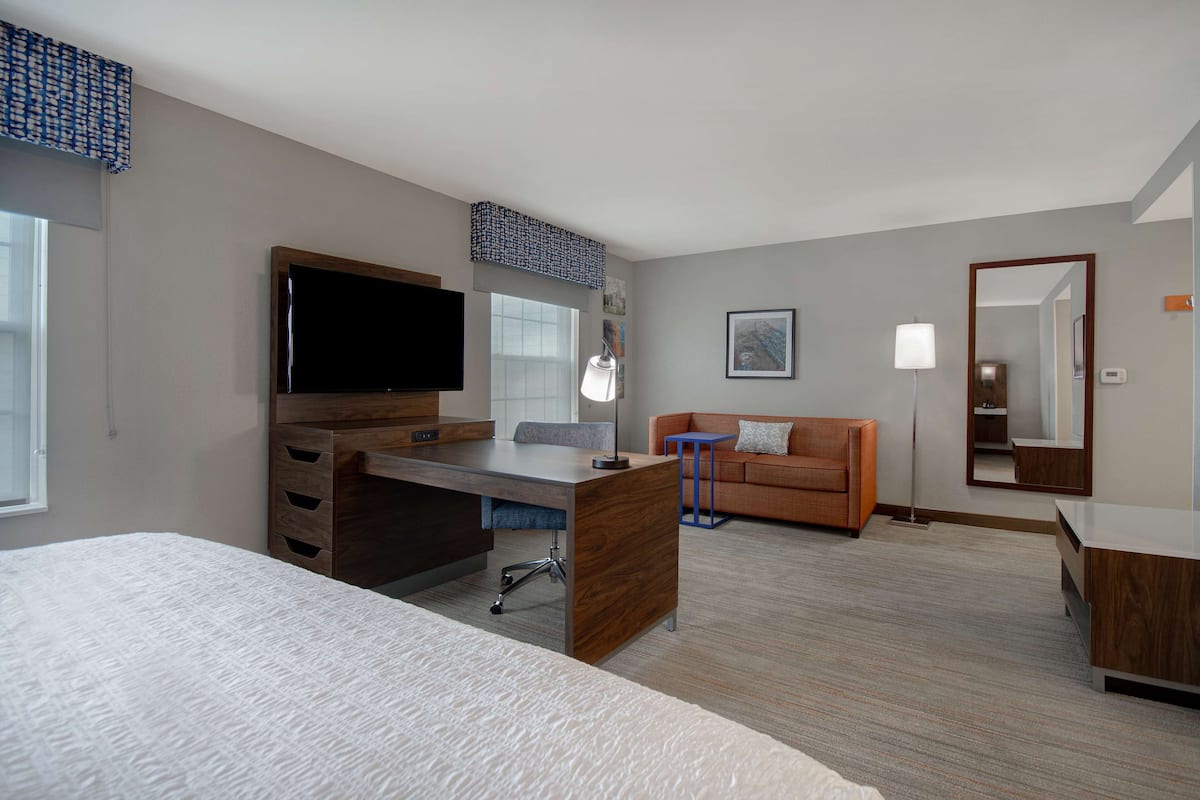 A hotel room with a bed, a desk with a flat-screen TV, a sofa, a small table, and a mirror.