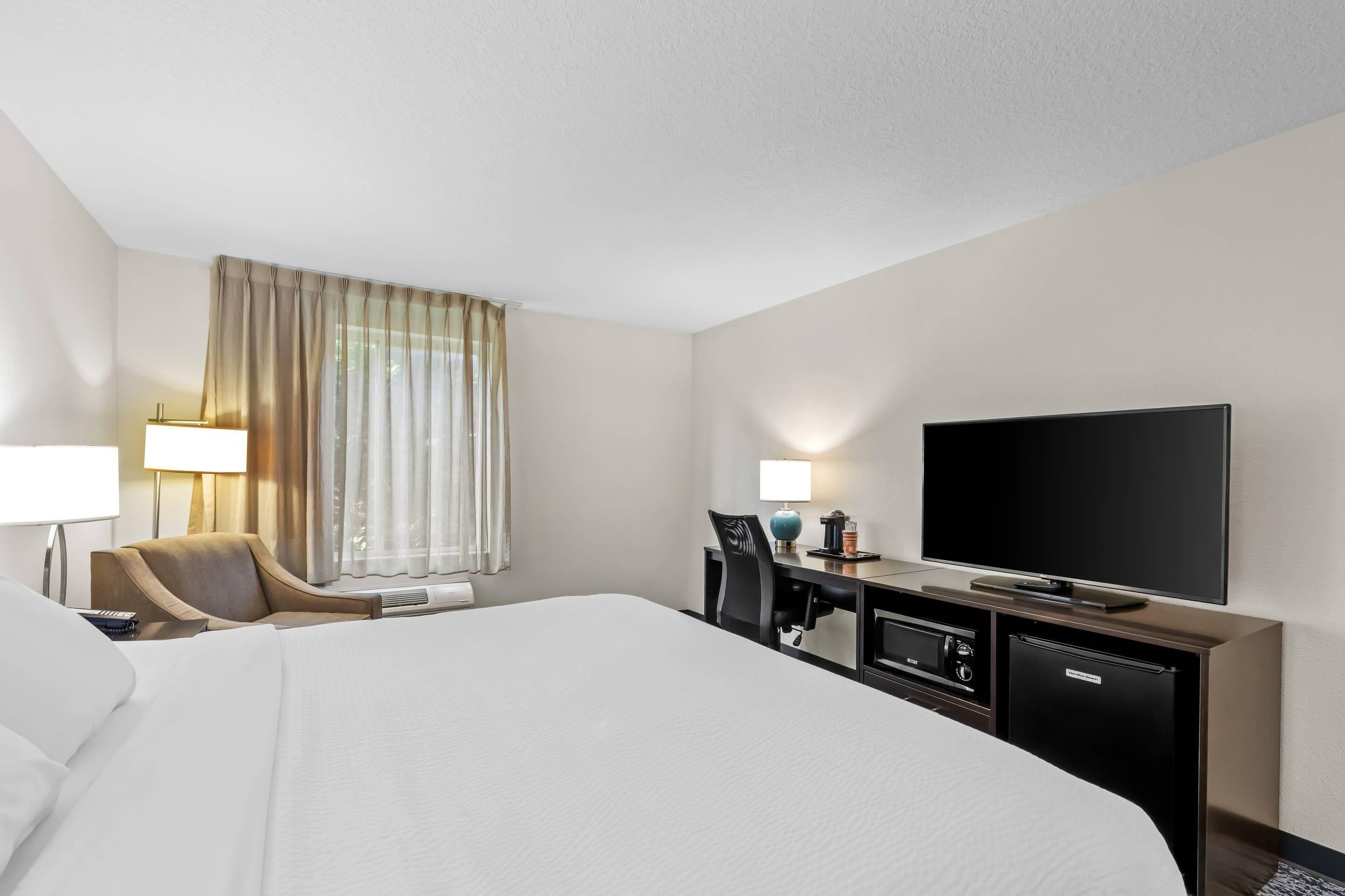 A hotel room with a large bed, a desk with a television, a chair, and a lamp.