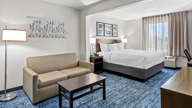 A hotel room with a bed, a sofa, a small table, a lamp, a picture on the wall, and a window with curtains. - Sleep Inn & Suites Hiram - Atlanta Northwest (Hiram)