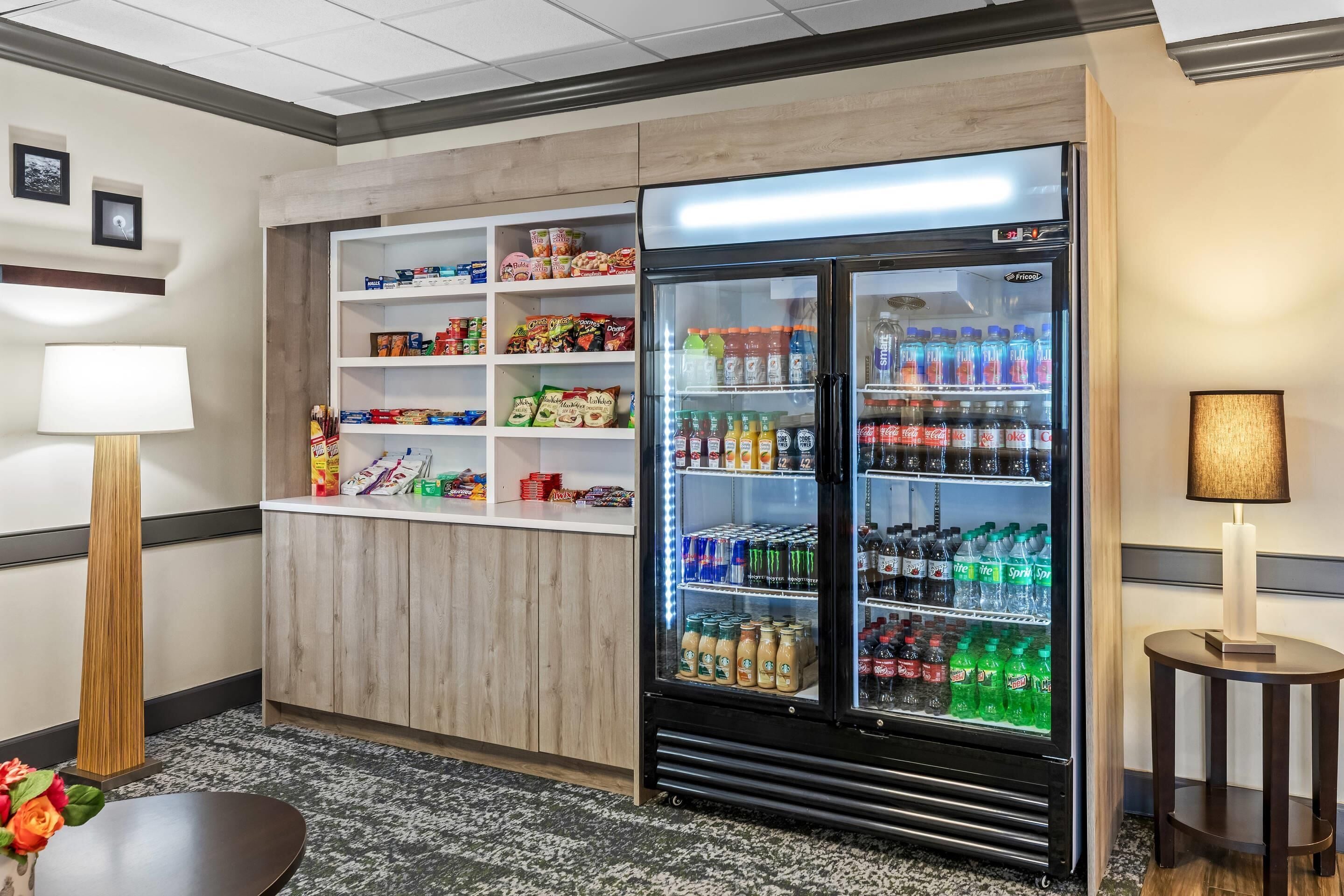 Habitación de hotel con minibar, refrigerador lleno de bebidas y una mesita con lámpara.