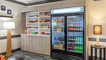Habitación de hotel con minibar, refrigerador lleno de bebidas y una mesita con lámpara.