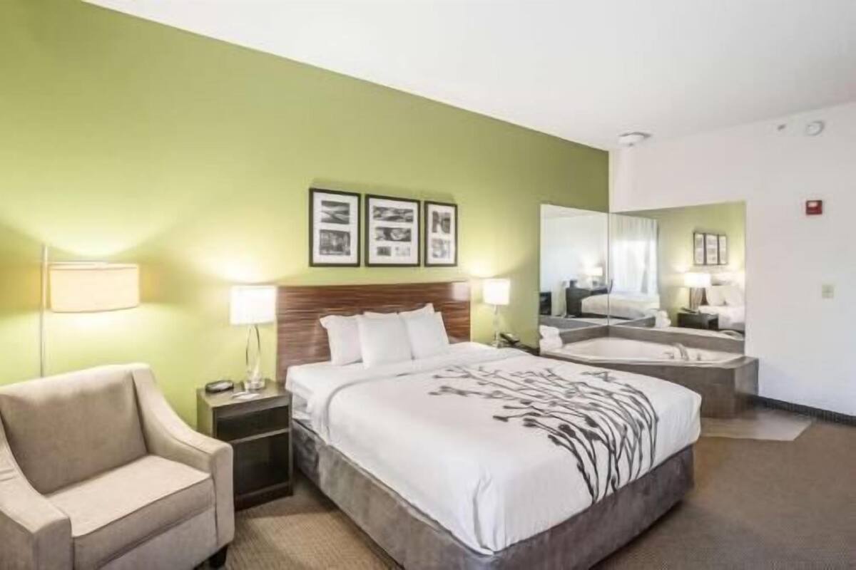 Habitación de hotel con cama, mesitas de noche, una silla, un espejo grande y un baño con bañera.