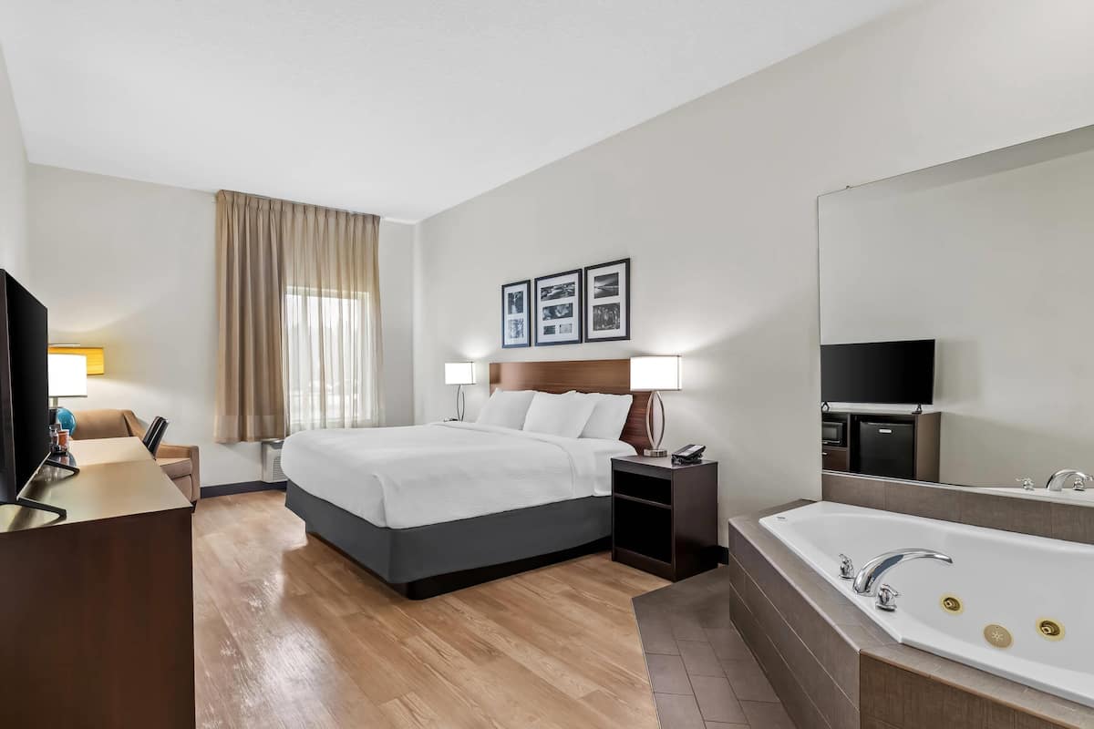 Una habitación de hotel con una cama grande, un escritorio con computadora, una lámpara de escritorio, una mesita de noche con teléfono, un espejo grande y una bañera.