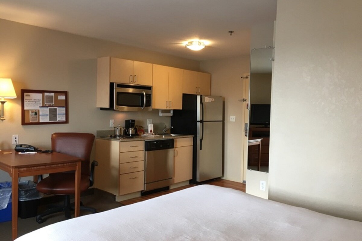 A compact hotel room with a bed, a small kitchenette including a microwave, refrigerator, and coffee maker, a desk with a chair, and a bulletin board on the wall.