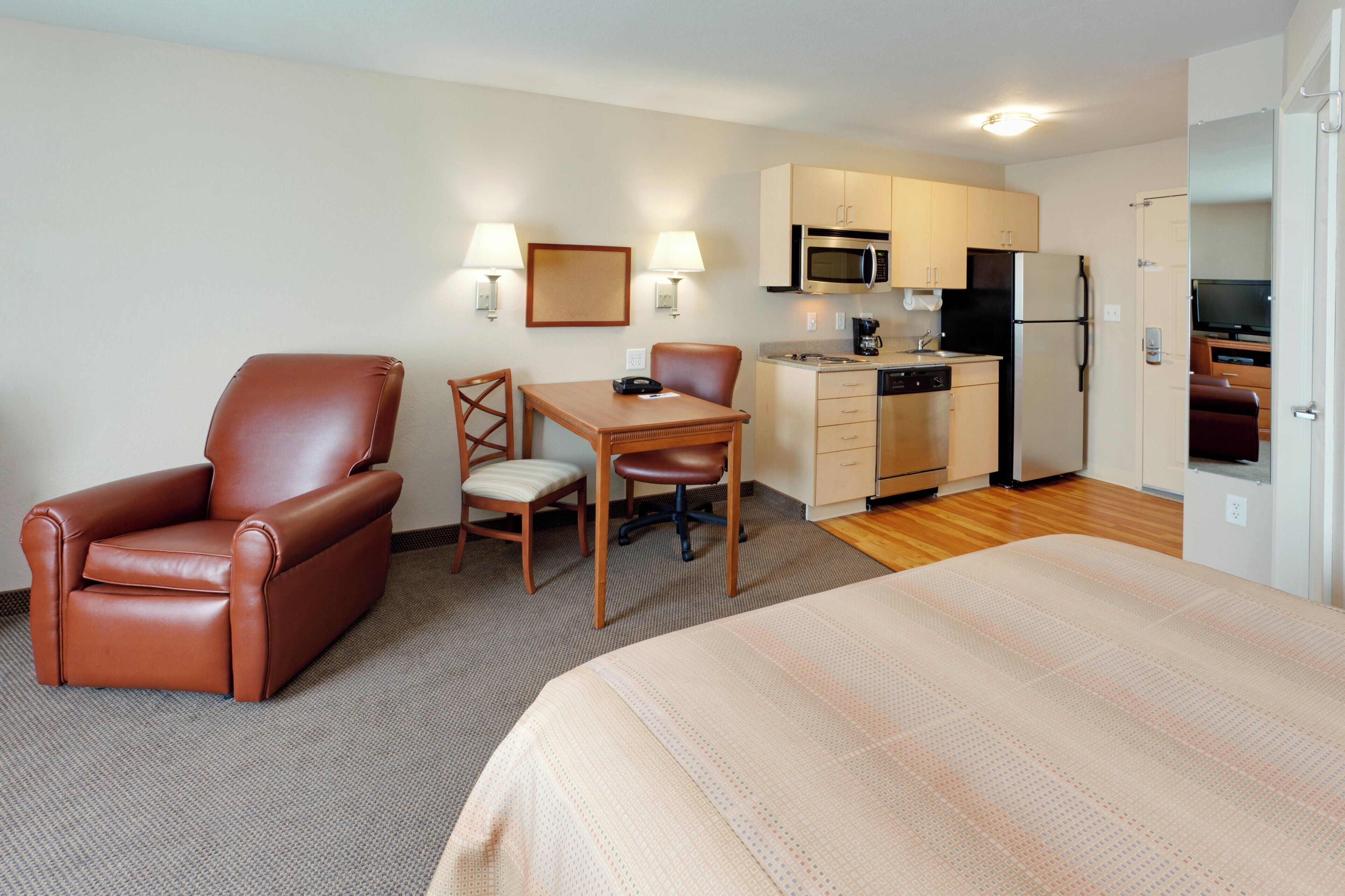 A hotel room with a bed, a desk, a chair, a kitchenette, and a television.