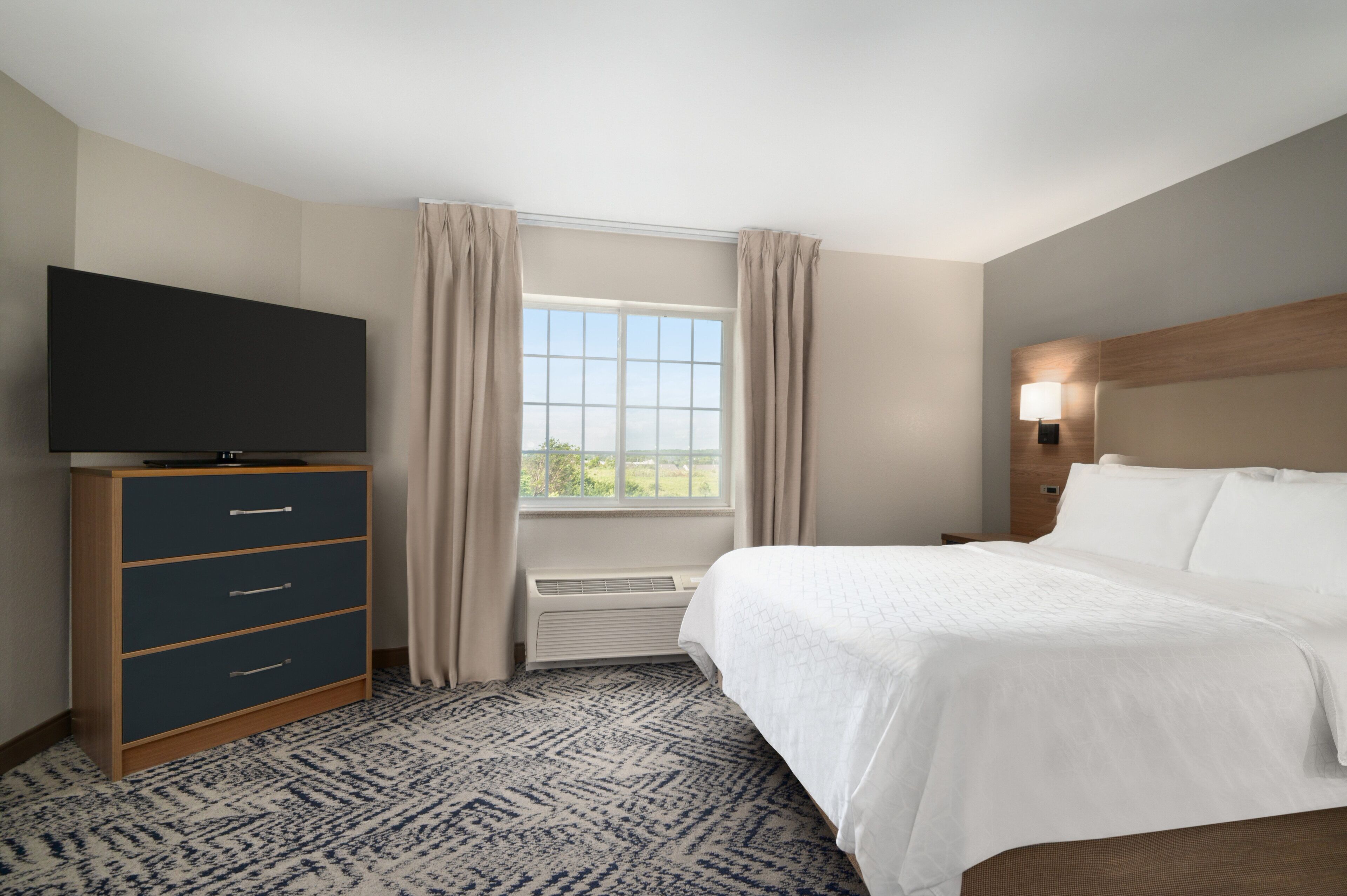 Hotelkamer met een bed, een tv op een standaard, een raam met gordijnen en een wandlamp.