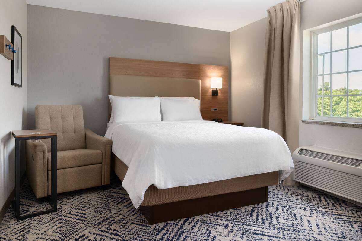 Habitación de hotel con cama, una silla, una mesita de noche, una lámpara y ventana con vistas.