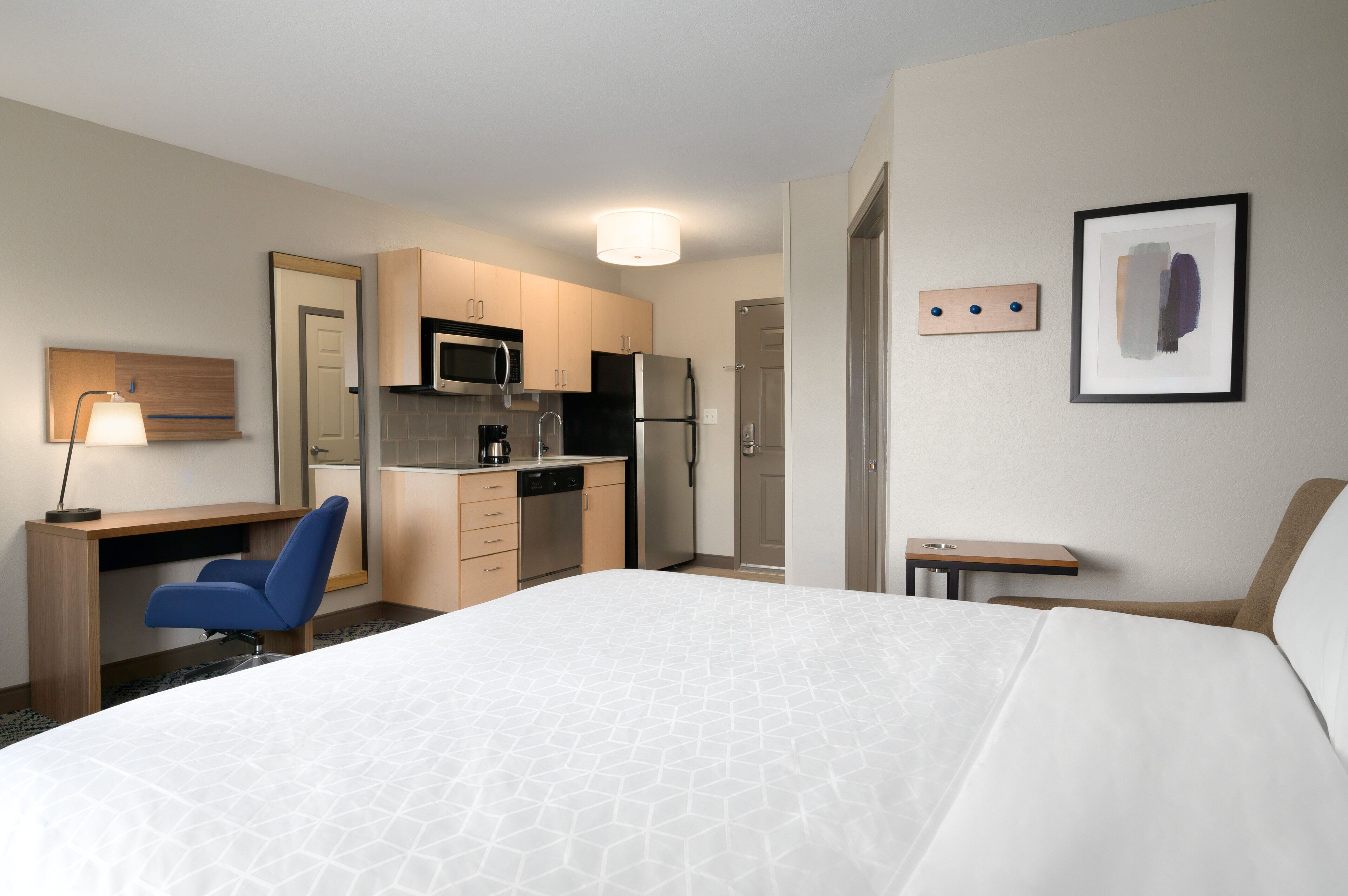 A hotel room with a bed, a desk, a chair, a kitchenette, and a small table.