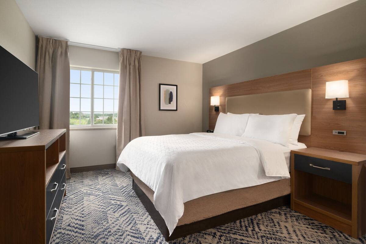 Habitación de hotel con cama, televisión, mesita de noche, lámpara y ventana con cortinas.