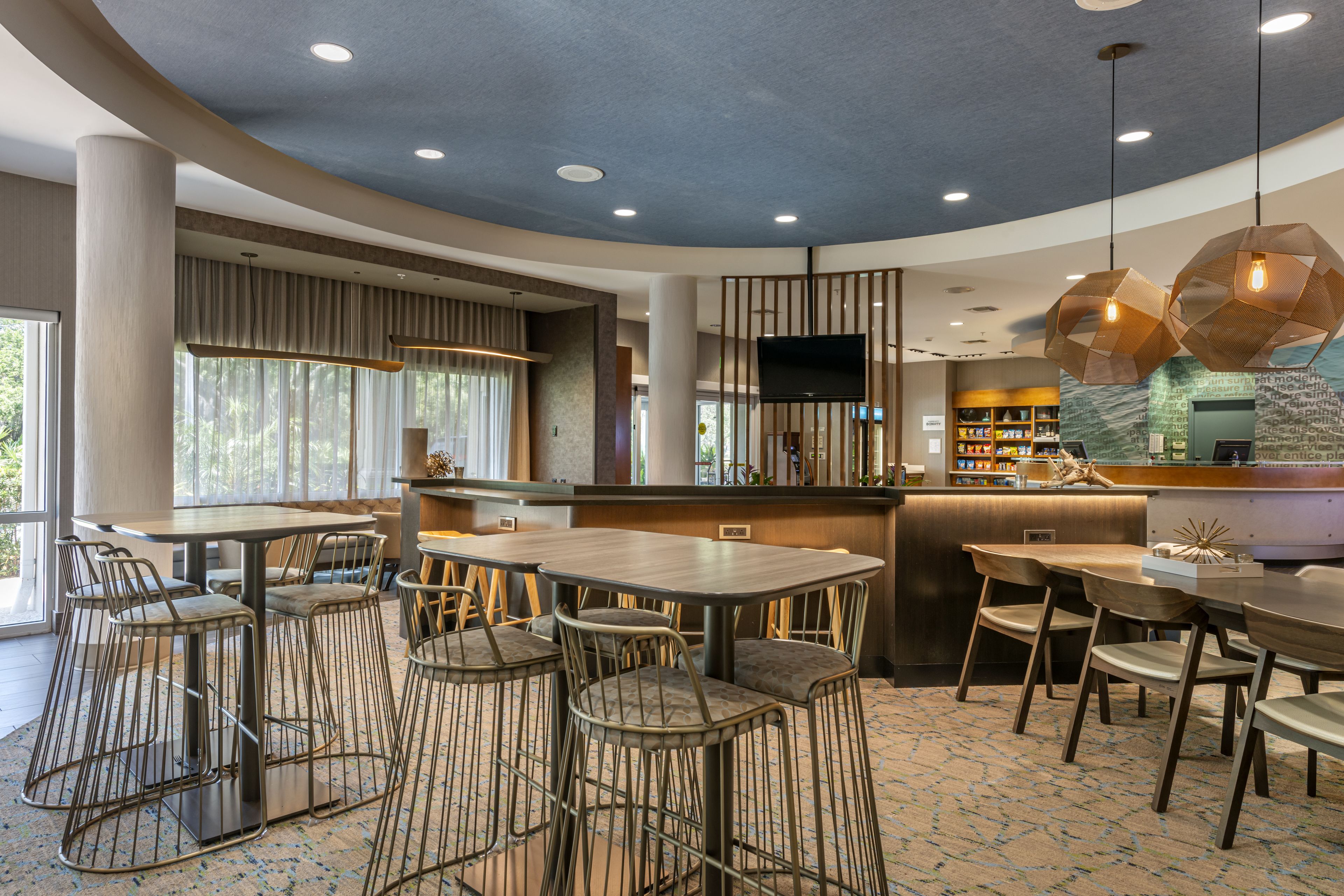A modern restaurant with high tables and metal bar stools, a bar area with a flat-screen TV, and a view of the outdoors.