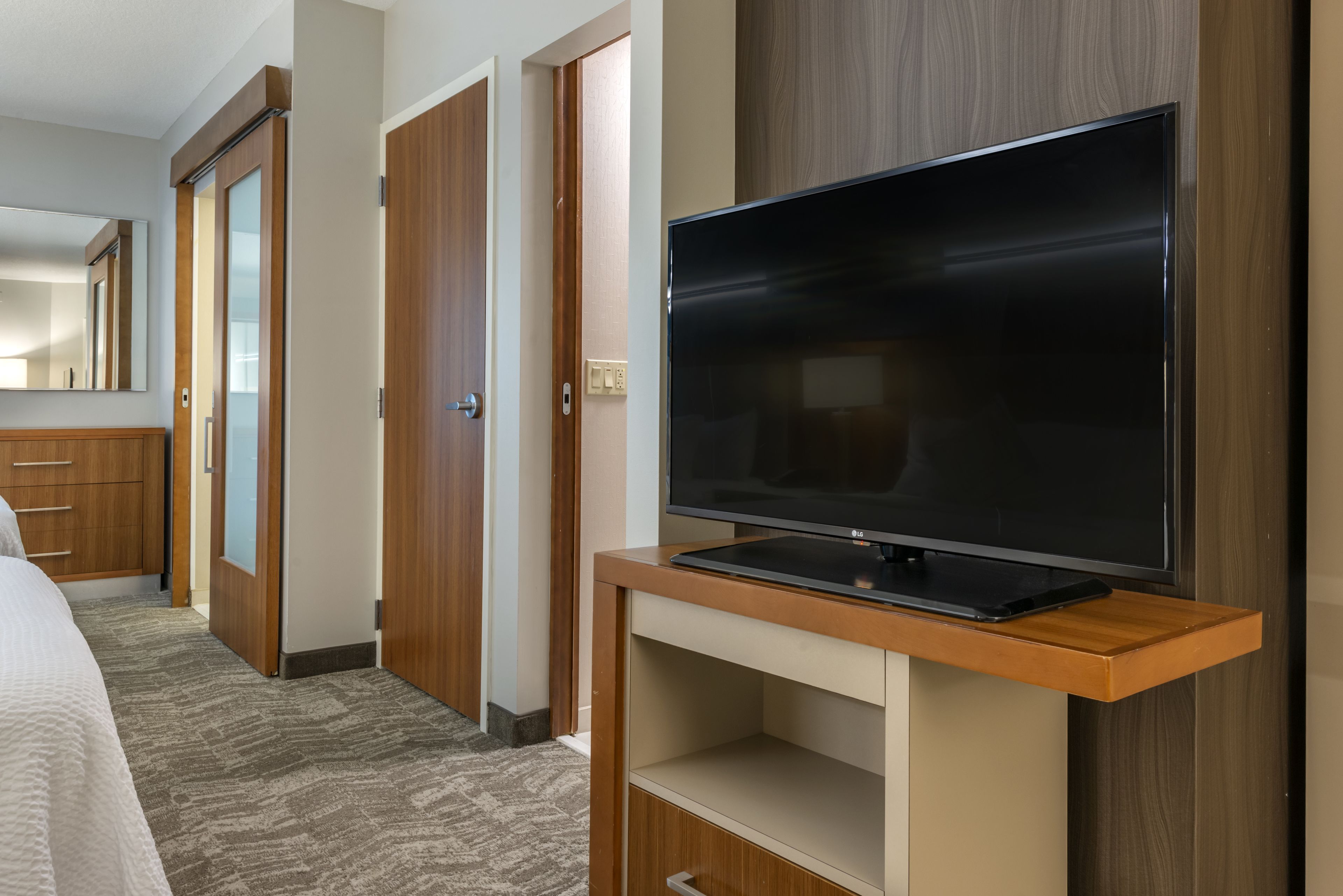 Habitación de hotel con un televisor de pantalla plana grande sobre un soporte de madera, una cama con cubrecama blanco, un armario de madera y un espejo en la pared.