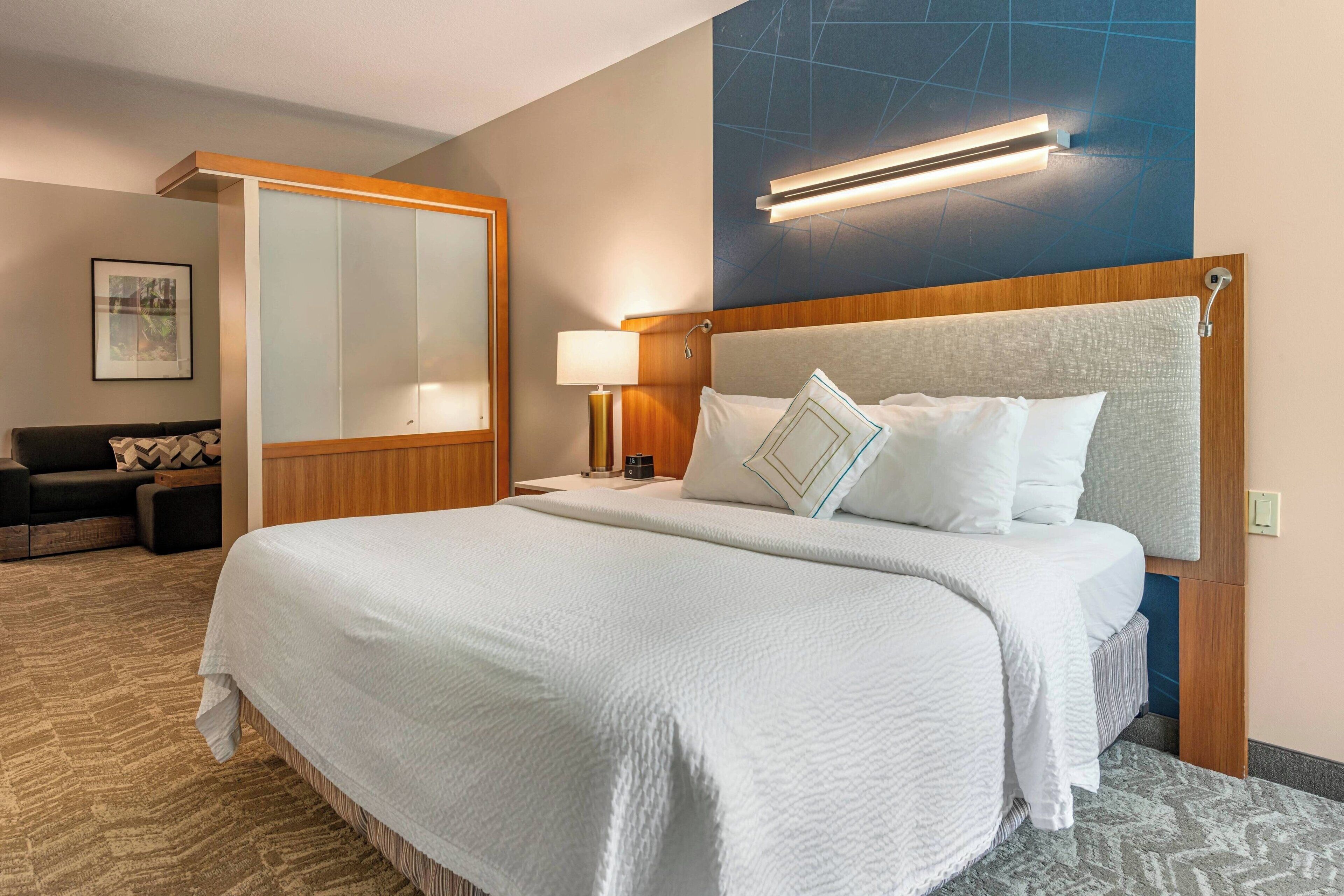 Una habitación de hotel con una cama grande, una mesita de noche con lámpara, un espacio de descanso con sofá y un espejo amplio.