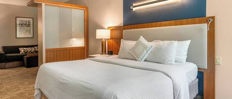 Una habitación de hotel con una cama grande, una mesita de noche con lámpara, un espacio de descanso con sofá y un espejo amplio.