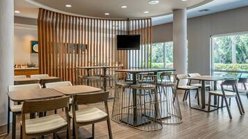 Zona pranzo moderna con tavoli e sedie in legno, TV a parete e ampie finestre con vista sul verde.