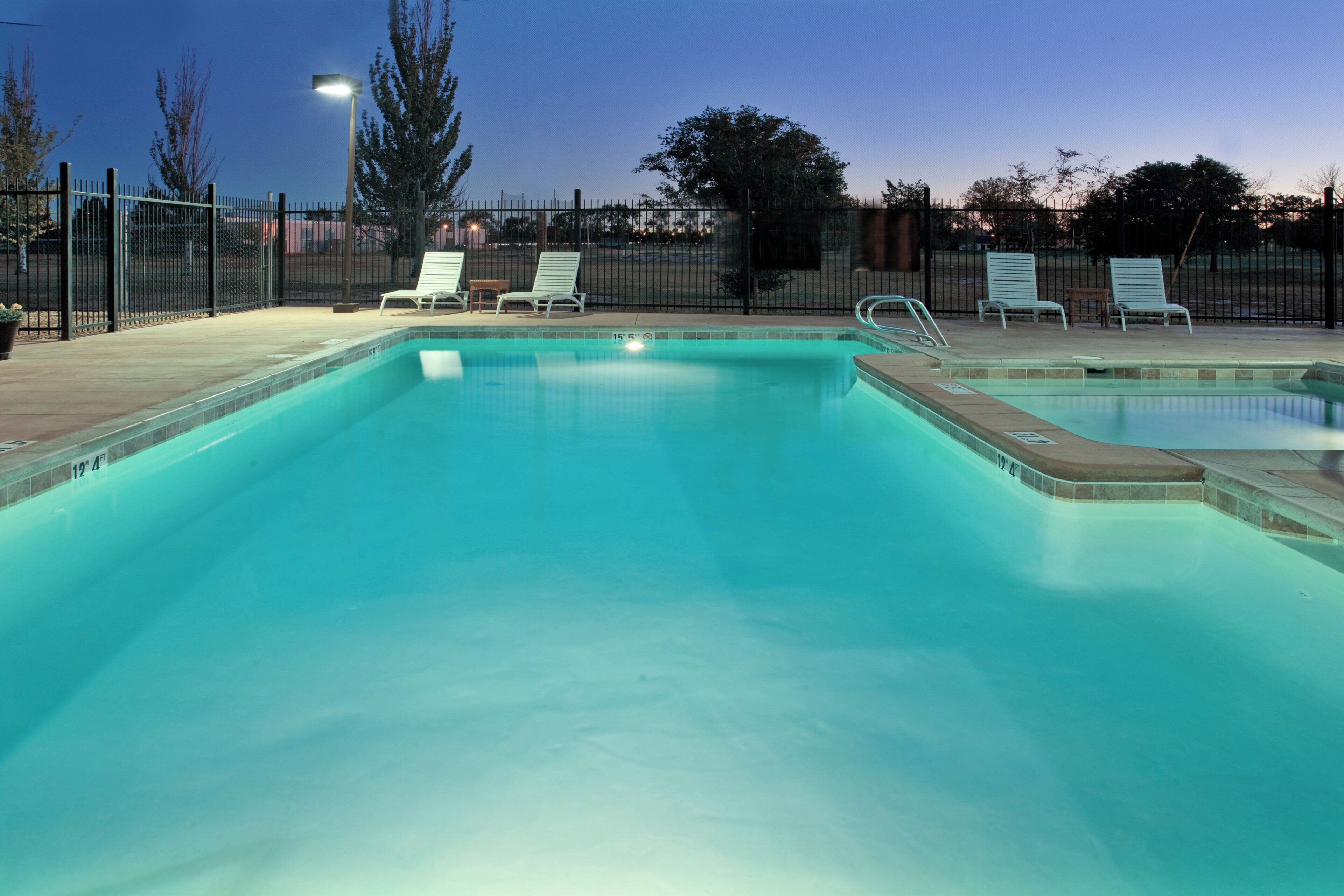 A rectangular swimming pool with a tiled edge, surrounded by a fence and several lounge chairs.