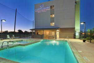 Exterior de un hotel con piscina, un letrero que dice 'CANDLEWOOD SUITES' y un área exterior cercada con sillas de descanso.