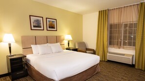 A hotel room with a large bed, two bedside tables with lamps, a chair, a window with curtains, and two framed pictures on the wall.