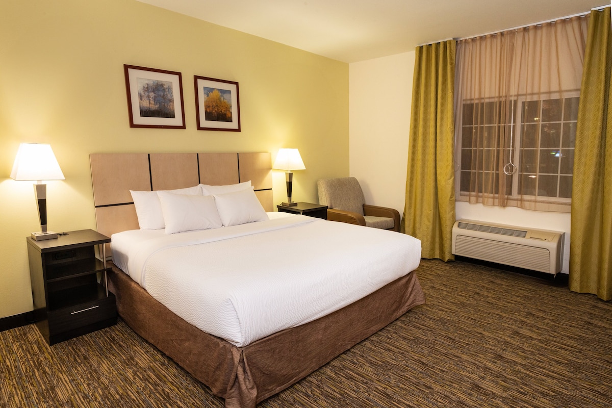 A hotel room with a large bed, two bedside tables with lamps, a chair, a window with curtains, and two framed pictures on the wall.