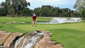 A golf course with a waterfall, a pond, and a golfer in a red shirt and beige shorts.