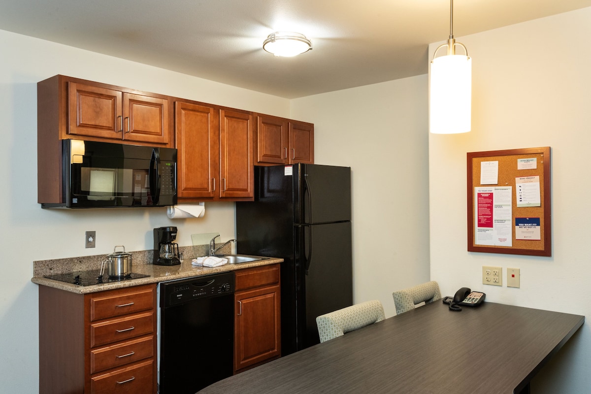 A kitchen with wooden cabinets, a black refrigerator, a microwave, a coffee maker, and a dishwasher. There is a bulletin board on the wall and a telephone on the table.