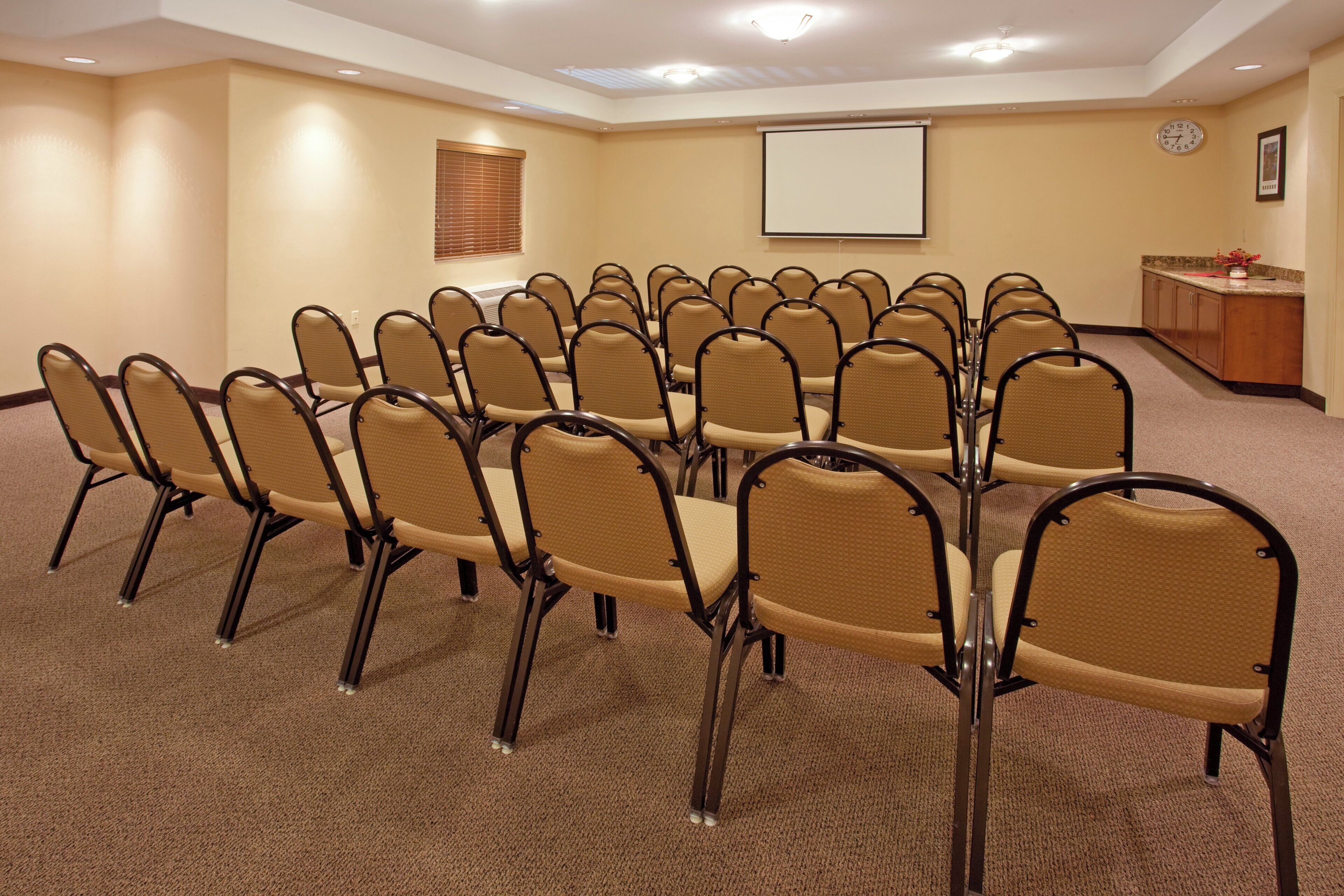 Un salón de conferencias con filas de sillas, una pantalla para proyector, un reloj y una pequeña área de cocina.